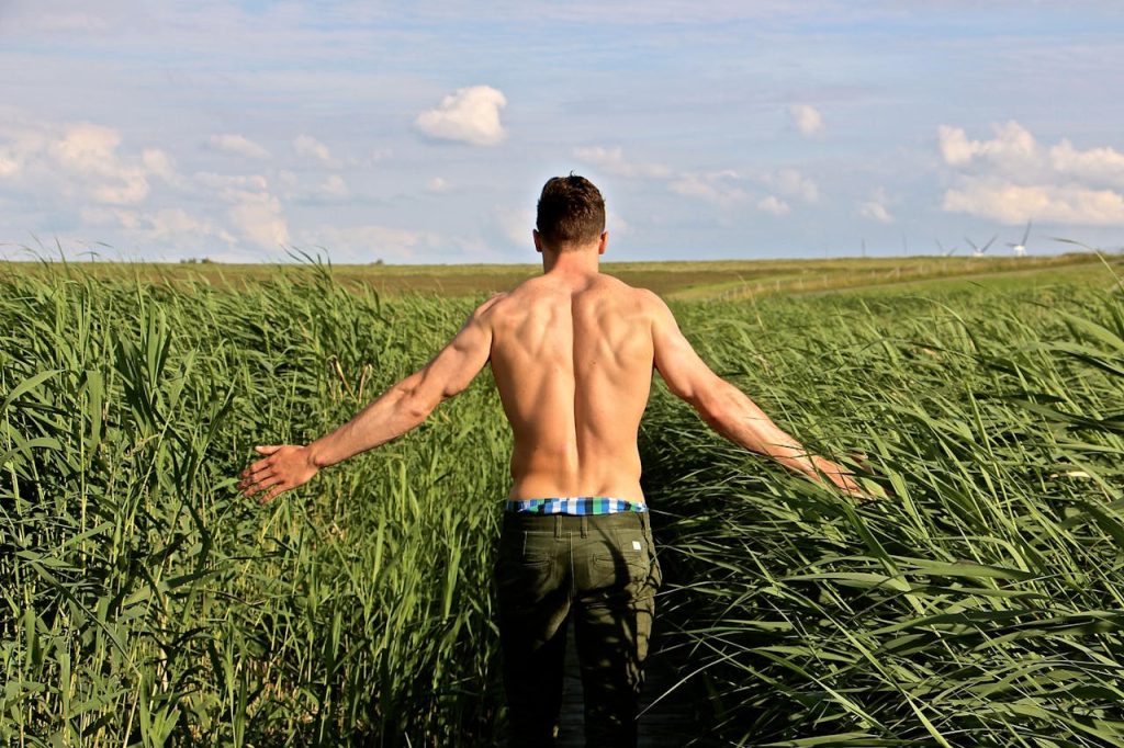 A muscular man walks through a lush green field, enjoying the windy countryside and scenic sky.
